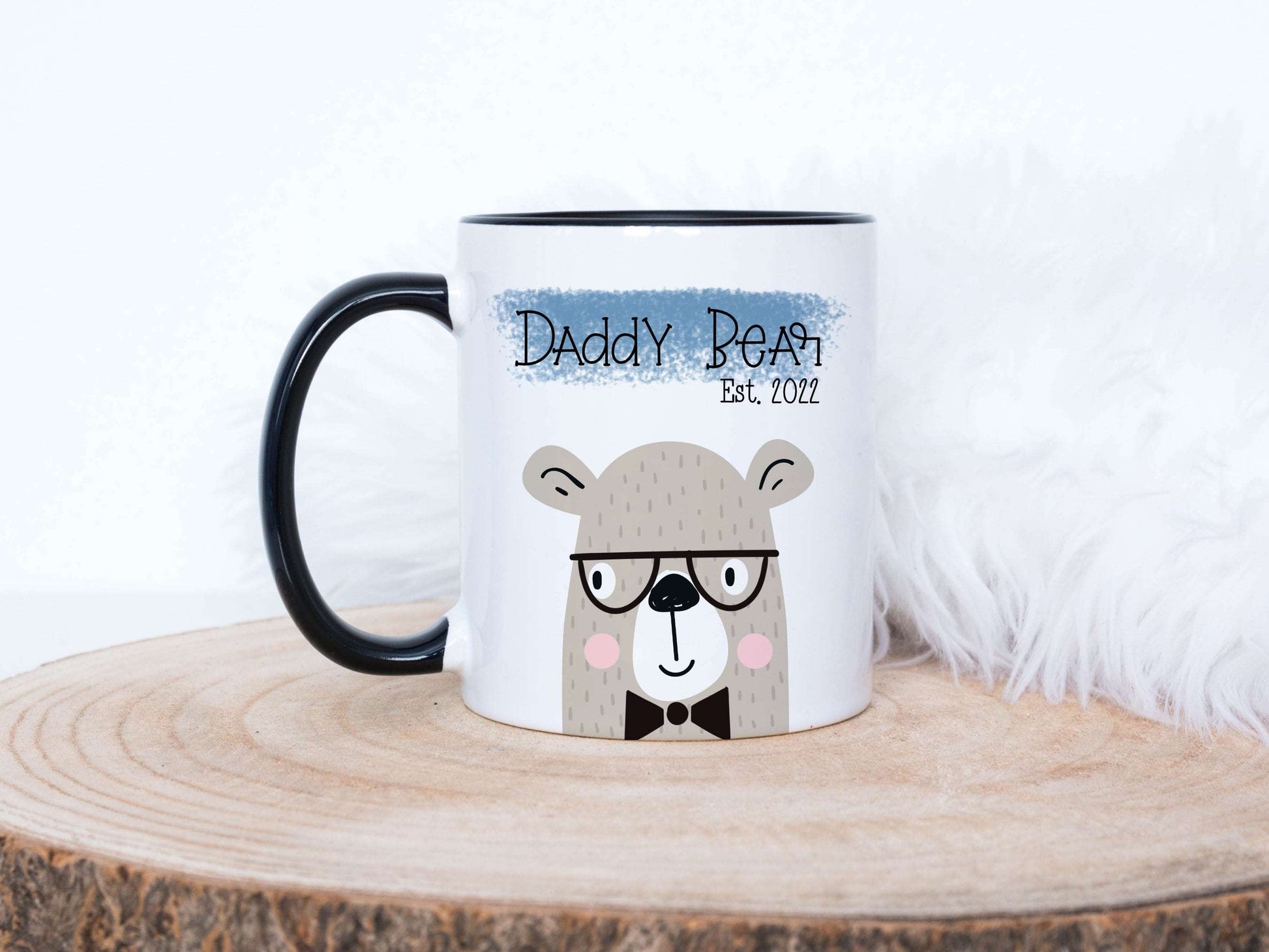 white mug with a cute bear design printed on with the text daddy bear above, The mug is sitting on a wood slice with fluffy background behind