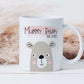 white mug with cute bear illustration printed on with the text mummy bear est. 2022 over a pink paint stroke effect smear. The mug is on a white wood effect surface with boho style grass behind