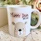 white mug with cute bear illustration printed on with the text mummy bear est. 2020 over a pink paint stroke effect smear. The mug is on a marble effect surface with flowers behind