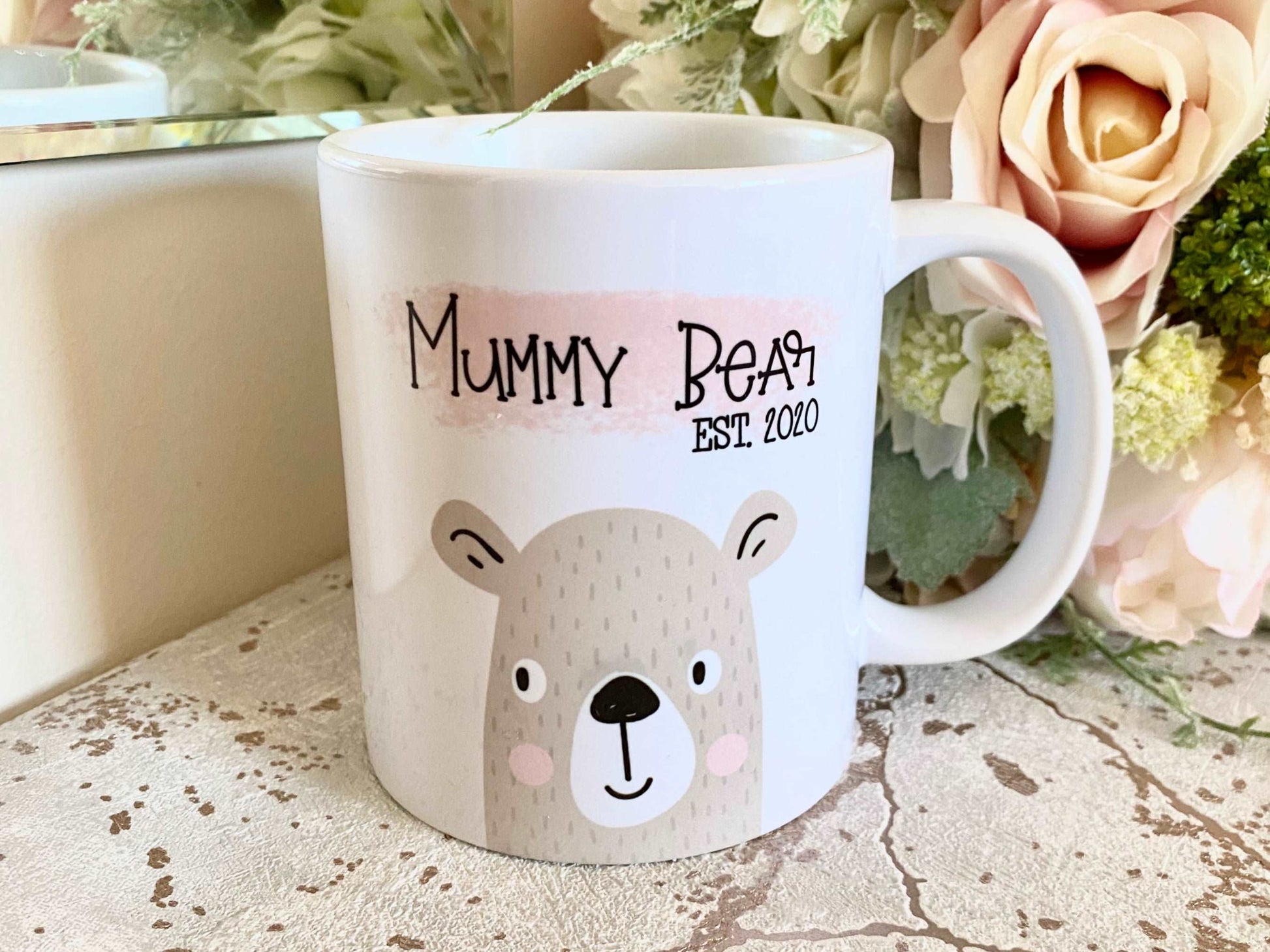 white mug with cute bear illustration printed on with the text mummy bear est. 2020 over a pink paint stroke effect smear. The mug is on a marble effect surface with flowers behind
