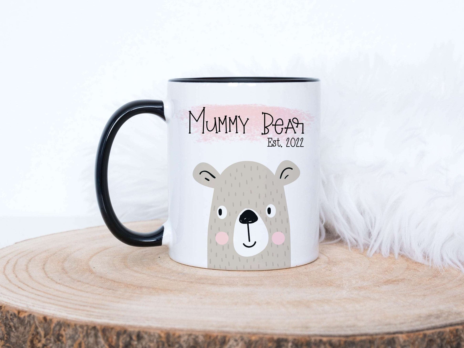 mummy bear design printed on a white mug with black handle. The mug is sitting on a wood slice in front of a white fluffy background