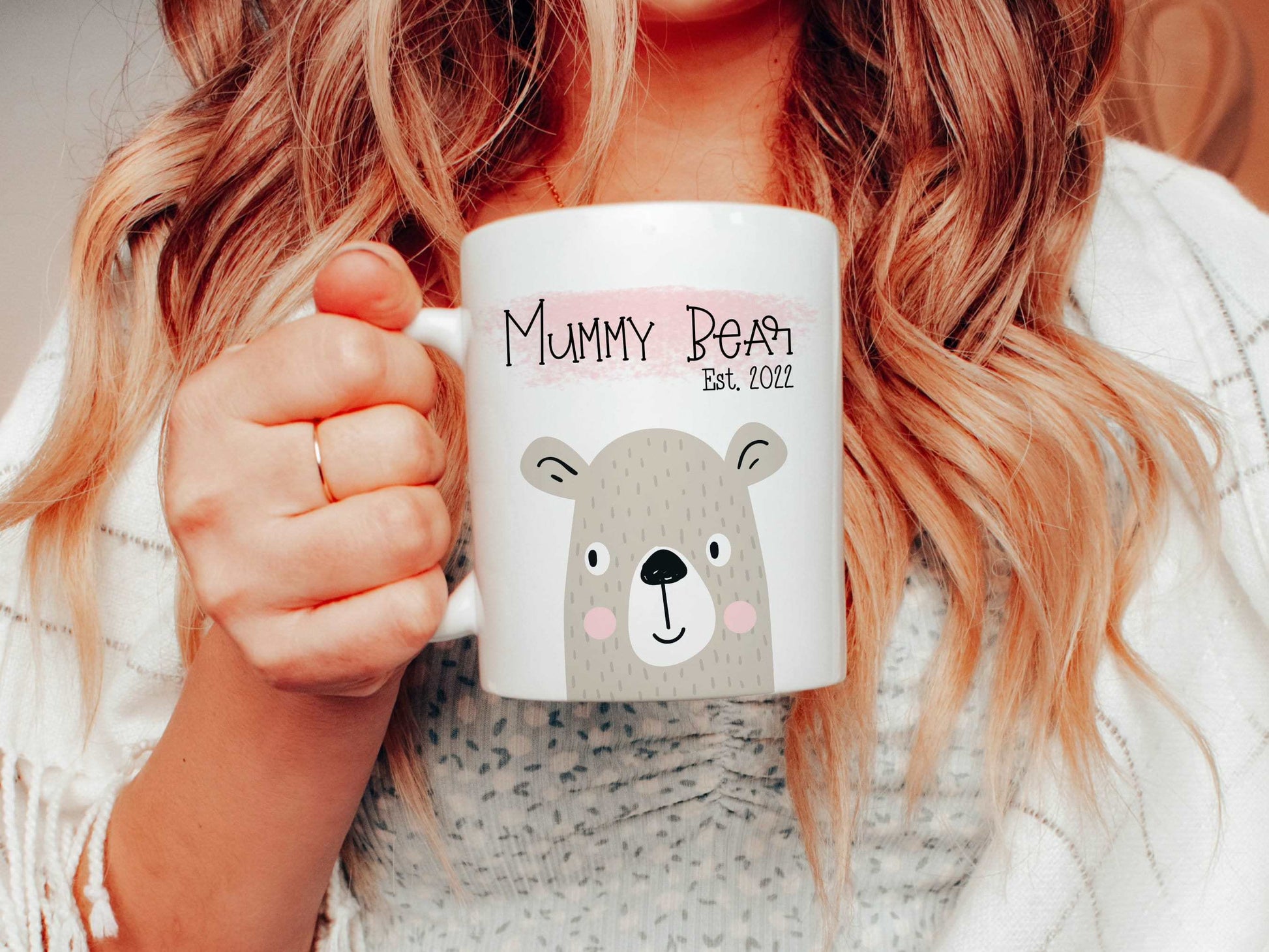 woman holding a white mug with a bear illustration on with the text mummy bear est. 2022 printed above over a pink brush stroke.
