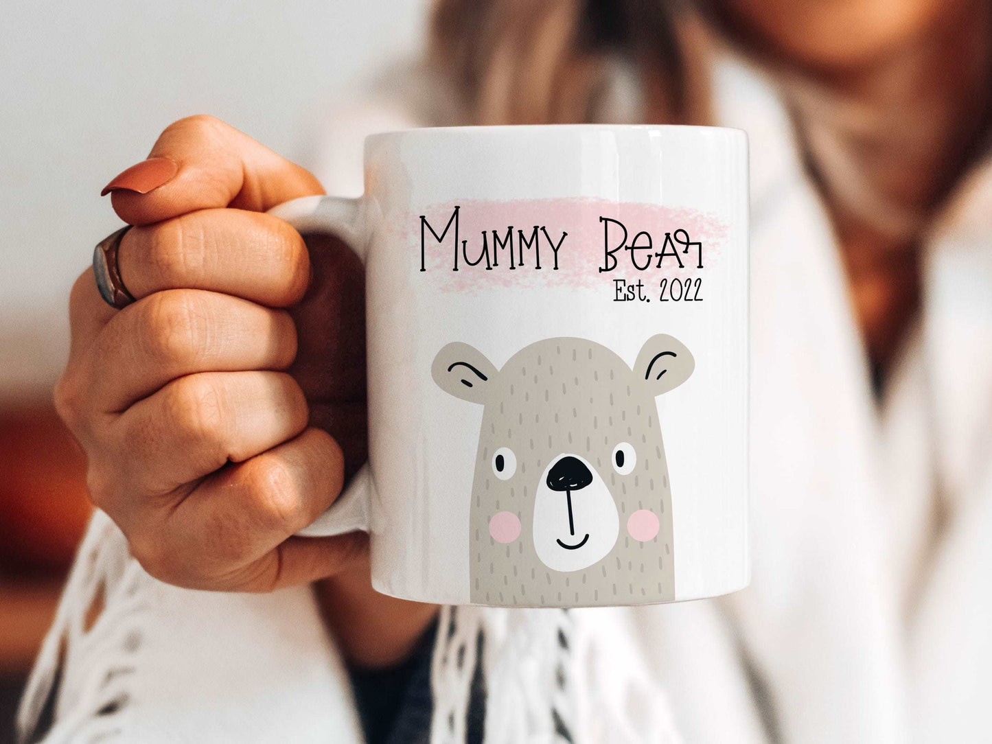 woman holding a white mug out in front of her with a bear illustration on with the text mummy bear est. 2022 printed above over a pink brush stroke.