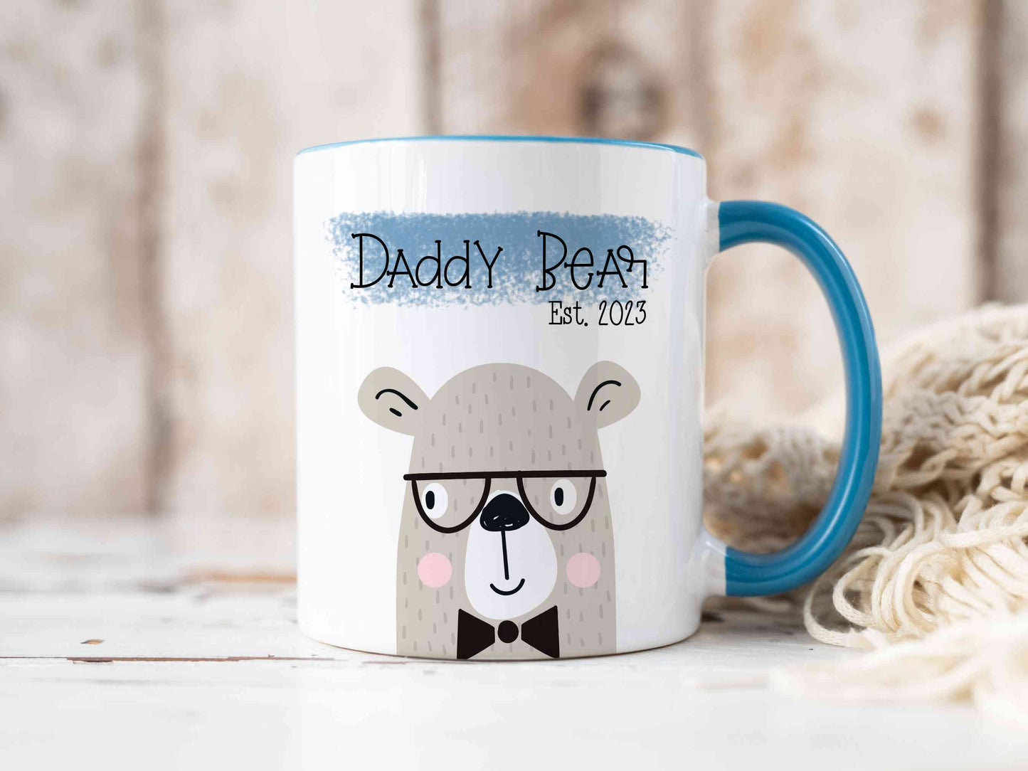 white mug with a blue handle sitting on a worktop. There is a cute bear design printed on the mug with the text daddy bear above over a blue banner.