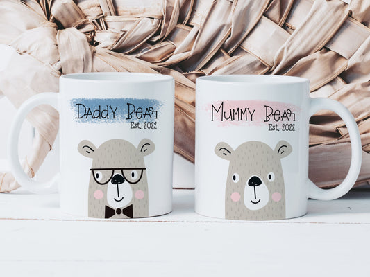 two white mugs on a white surface. One mug has the text mummy bear est 2022 over a pink paint stroke, the other has daddy bear est 2022 over a blue paint stroke. Both have a cute bear illustration printed below the text
