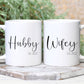 two white mugs on a tile with greenery behind. One mug has the text hubby in a script font with est. 2022 below. The other has wifey, also with est 2022 below.