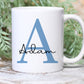 white mug with greenery behind. The mug has a large blue letter A printed with the name Adam over the top in a black script font