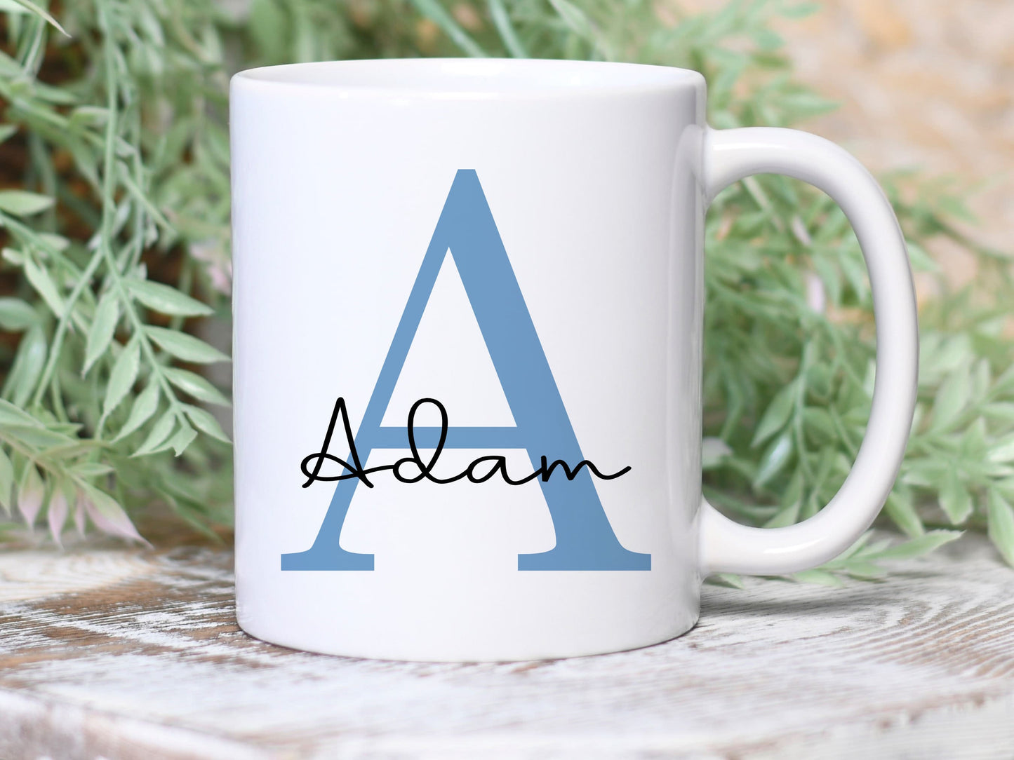 white mug with greenery behind. The mug has a large blue letter A printed with the name Adam over the top in a black script font
