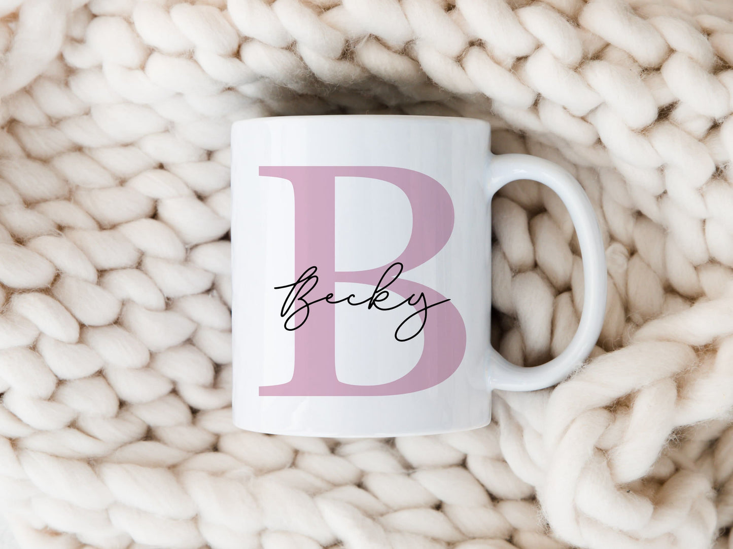 personalised pink letter mug with the name Becky printed over in a script font. The mug is on a cream knitted throw