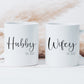 two white mugs with a boho style grass behind. One mug has the text hubby printed on in a script font and smaller text below stating est. 2022. The other mug is similar with the word wifey on.