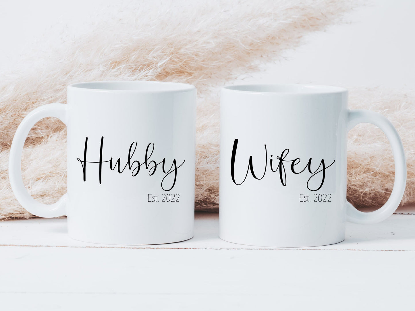 two white mugs with a boho style grass behind. One mug has the text hubby printed on in a script font and smaller text below stating est. 2022. The other mug is similar with the word wifey on.