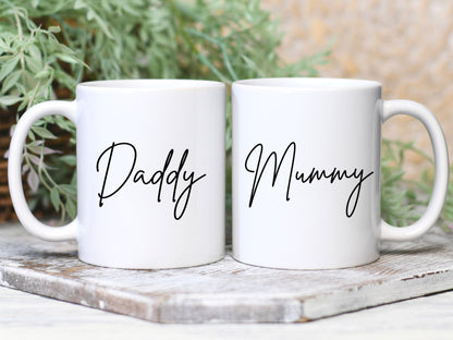 two white mugs on a tile with greenery behind. One has the text daddy printed on in a script font, the other has mummy printed on